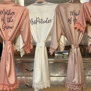 May include: Four satin robes with lace trim on the sleeves and hem. Three robes are dusty rose, and one is white. The dusty rose robes feature black text reading Mother of the Groom, Maid of Honor, and Ava. The white robe reads MrsPetrides.