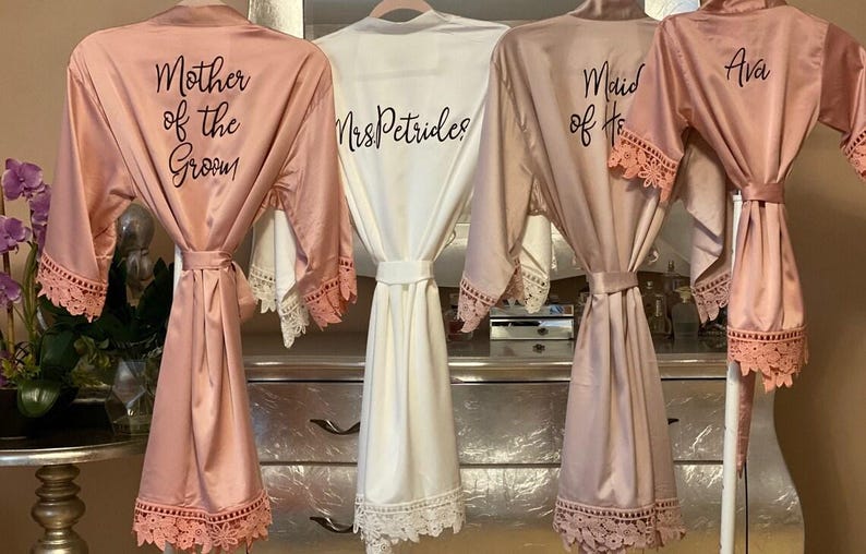 May include: Four satin robes with lace trim on the sleeves and hem. Three robes are dusty rose, and one is white. The dusty rose robes feature black text reading Mother of the Groom, Maid of Honor, and Ava. The white robe reads MrsPetrides.