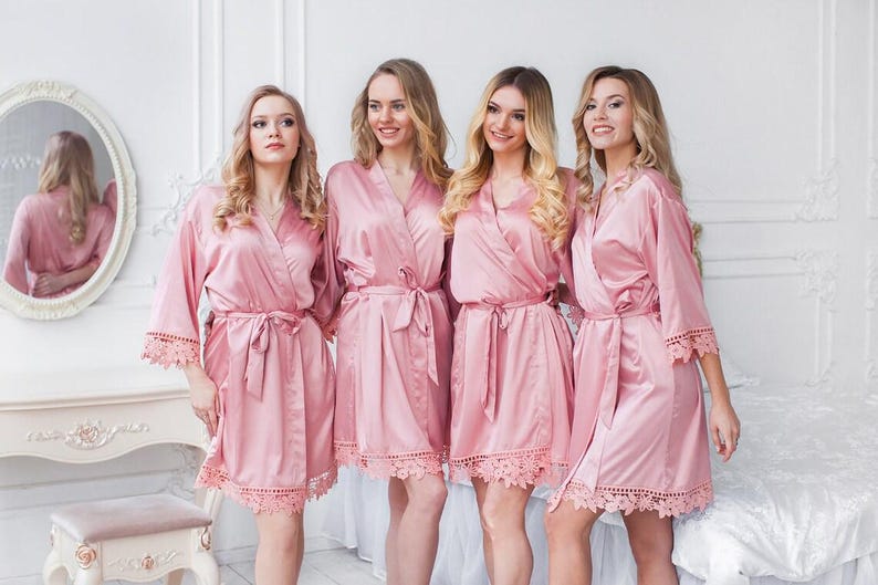 May include: Four matching dusty rose satin robes featuring long sleeves, a wrap-front design with a tie waist, and intricate lace trim detailing along the sleeve cuffs and the bottom hemline of each garment, creating a coordinated set.