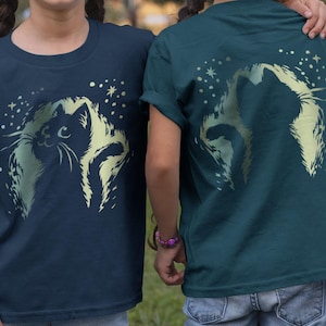 Könnte beinhalten: Zwei Kinder tragen marineblaue und teal-farbene T-Shirts mit einem Katzenmotiv. Die Vorderseite des marineblauen Shirts und die Rückseite des teal-farbenen Shirts zeigen ein Katzendesign mit Sternen. Beide Kinder tragen blaue Jeans.