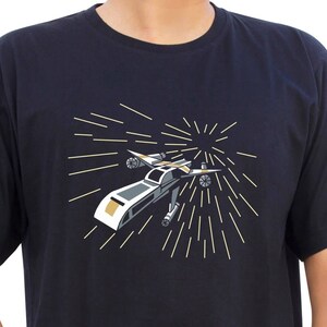 Könnte beinhalten: Ein schwarzes T-Shirt mit einer weißen und goldenen Grafik eines Raumschiffs, das durch den Weltraum fliegt. Das Raumschiff ist von einem Sternenausbruchseffekt umgeben.