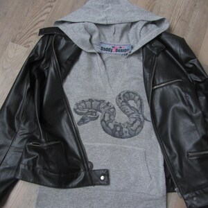 Könnte beinhalten: Ein grauer Kapuzenpullover mit einem schwarzen Schlangenmotiv und dem Text "Daddy Design" auf der Vorderseite. Der Pullover ist über eine schwarze Lederjacke gelegt.