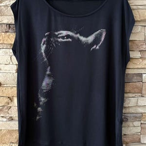May include: Black scoop-neck t-shirt with a silver dotted design of a face. The short-sleeved top is hanging on a black hanger against a light brown brick wall. The bottom hem is slightly longer in the back.