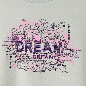 Puede incluir: Camiseta azul claro con un estampado gráfico de la palabra "DREAM" en letras moradas y rosas. Las letras están rodeadas de un diseño de letras y símbolos más pequeños.