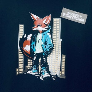 Könnte beinhalten: Schwarzer Hoodie mit einem Grafikdruck eines Fuchses, der eine blaue Jacke trägt und vor einer Skyline steht. Der Text "Lehtestefir Daddy2Design" ist auf einem weißen Etikett gedruckt.