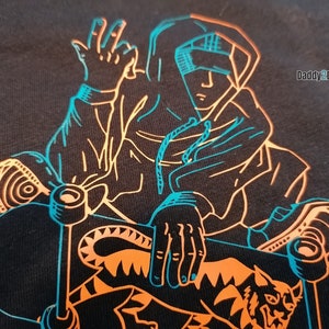 Puede incluir: Una camiseta negra con un diseño gráfico de una persona haciendo skateboard. La persona lleva una sudadera con capucha y tiene el brazo levantado en el aire. El gráfico está delineado en naranja y azul. El texto "Daddy2Design" está en la esquina superior derecha de la imagen.