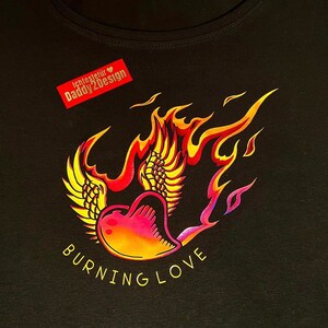 Könnte beinhalten: Ein schwarzes T-Shirt mit einem Grafikdruck eines Herzens in Flammen mit Flügeln und dem Text "BURNING LOVE". Die Grafik ist in Gelb-, Orange-, Rot- und Lilatönen gehalten. Der Text "lichtestijl Daddy2Design" befindet sich in einem roten Kasten mit einem Herzsymbol.