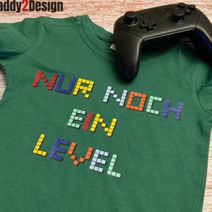 Könnte beinhalten: Grünes Langarmshirt mit dem Text "NUR NOCH EIN LEVEL" in bunten Pixelbuchstaben.