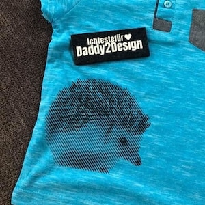 May include: A turquoise blue t-shirt with a black and white hedgehog graphic. The shirt has a small pocket on the left side and a button-up collar. The text "Ichtestefür Daddy2Design" is on a black label.