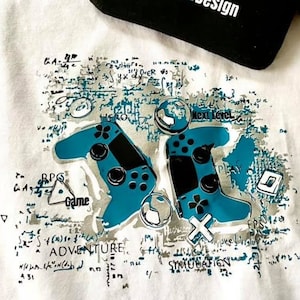Puede incluir: Camiseta blanca de manga larga con un estampado gráfico de dos mandos de videojuegos azules y negros. El gráfico también incluye las palabras "Next Level", "Game", "Adventure" y "Simulation". La camiseta tiene una bolsa negra con cremallera con el texto "Ichlestiefür Daddy2Design" en ella.