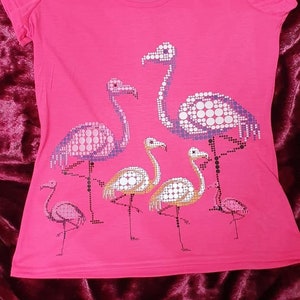 May include: A bright pink t-shirt featuring a flamingo design. The design includes several flamingos in various sizes, with dot patterns in white, purple, and orange. The brand name "Janino" is printed on the top.