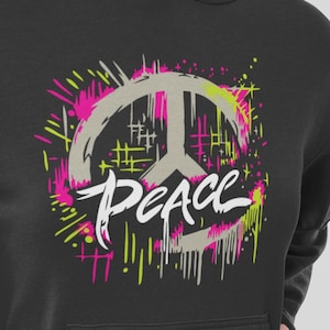 May include: Black hoodie featuring a peace sign graphic with the word "Peace" in white script. The peace sign is gray, surrounded by pink and yellow paint splatter designs. A casual, comfortable style.