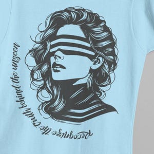 Könnte beinhalten: Hellblaues T-Shirt mit einer schwarz-weißen Illustration einer Frau mit Augenbinde. Das Design enthält den Text "Erkenne die Wahrheit hinter dem Unsichtbaren" in einer Schriftart. Das T-Shirt wird von zwei Händen gehalten.
