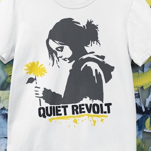 May include: A white short-sleeved t-shirt featuring a dark gray graphic of a person with a ponytail holding a bright yellow flower. Below the graphic, the text reads "QUIET REVOLT" in bold black letters with a yellow paint-splatter accent underneath.
