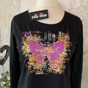 Op de afbeelding: Zwart longsleeve shirt met een paarse vlinderprint op een gouden en zwarte achtergrond. Het shirt heeft de tekst "Ich trage f&uuml;r Daddy2Design" op een rood label.