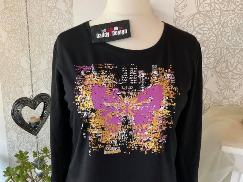 Op de afbeelding: Zwart longsleeve shirt met een paarse vlinderprint op een gouden en zwarte achtergrond. Het shirt heeft de tekst "Ich trage f&uuml;r Daddy2Design" op een rood label.