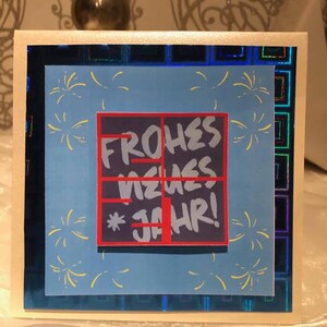 Puede incluir: Una tarjeta azul con un cuadrado rojo y negro con el texto "FROHES NEUES JAHR!" en blanco. El cuadrado est&aacute; rodeado de fuegos artificiales amarillos sobre un fondo azul.