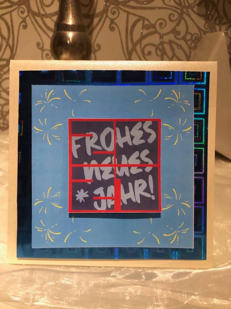 Puede incluir: Una tarjeta azul con un cuadrado rojo y negro con el texto "FROHES NEUES JAHR!" en blanco. El cuadrado est&aacute; rodeado de fuegos artificiales amarillos sobre un fondo azul.