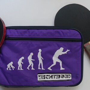 May include: Purple table tennis bag with a zipper closure. The bag features a white silhouette of the evolution of humans playing table tennis. The text "TISCHTENNS" is printed on the bag. The bag is next to a black table tennis paddle with a wooden handle.