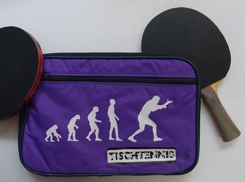 May include: Purple table tennis bag with a zipper closure. The bag features a white silhouette of the evolution of humans playing table tennis. The text "TISCHTENNS" is printed on the bag. The bag is next to a black table tennis paddle with a wooden handle.