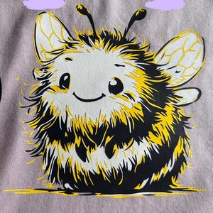 Puede incluir: Primer plano de una camiseta de color púrpura claro con un diseño de abeja de dibujos animados. La abeja tiene una cara blanca, ojos negros y una boca sonriente. Su cuerpo es negro y amarillo, con alas amarillas. Dos formas de nube púrpura están encima de la abeja.