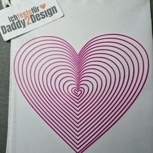 May include: A white drawstring bag with a large, concentric heart design in shades of pink. The bag has a tag that reads "Ich teste für Daddy2Design."