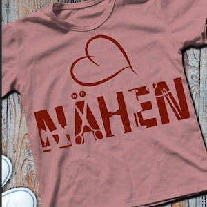 Può includere: Una maglietta rosa con un cuore rosso e la parola "NÄHEN" in lettere rosse. La parola è composta da icone legate alla cucitura, tra cui un ago, filo, forbici e bottoni.