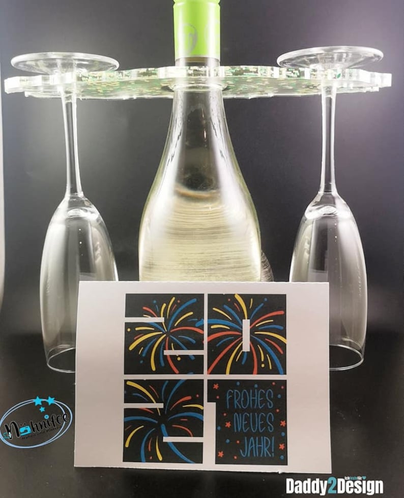 Puede incluir: Una botella de vino blanco con una tapa verde, dos copas de vino transparentes y un soporte para botellas de vino de acr&iacute;lico transparente. Una tarjeta con fondo negro y un gr&aacute;fico de fuegos artificiales coloridos dice "Frohes Neues Jahr!"