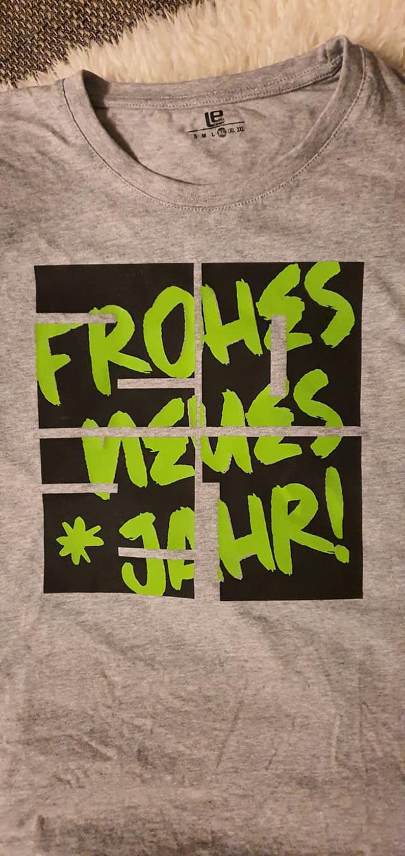 Puede incluir: Una camiseta gris con un gr&aacute;fico negro y verde que dice "Frohes Neues Jahr!"
