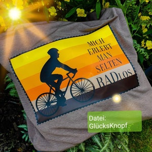 Könnte beinhalten: Ein graues T-Shirt mit einem Aufdruck einer Person, die Fahrrad fährt. Der Aufdruck ist im Retro-Stil mit orangefarbenen, gelben und braunen Streifen gehalten. Der Text auf dem Aufdruck lautet "Mich erlebt man selten radlos". Der Text "Datei: Glücksknopf" ist unter dem Aufdruck gedruckt.