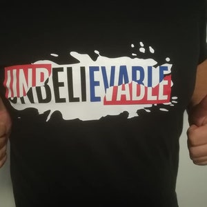 Könnte beinhalten: Ein schwarzes T-Shirt mit einem weißen Spritzmuster. Das Wort "UNGLAUBLICH" ist in roten, blauen und weißen Buchstaben über dem Design geschrieben.