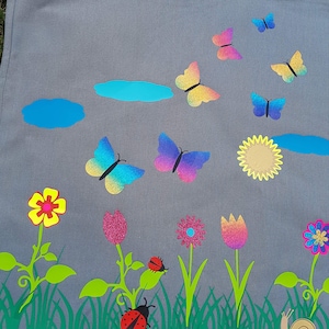 Könnte beinhalten: Eine graue Stofftasche mit einem bunten Schmetterlings-, Blumen- und Marienkäfer-Design. Die Tasche hat einen blauen Himmel, einen blauen Teich und eine gelbe Sonne.