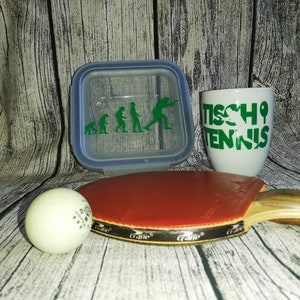 May include: A white ceramic mug with green text that reads "Tischtennis" and a green silhouette of a person playing table tennis. A white table tennis ball, a red and black table tennis paddle, and a clear plastic container with a green silhouette of the evolution of humans playing table tennis.