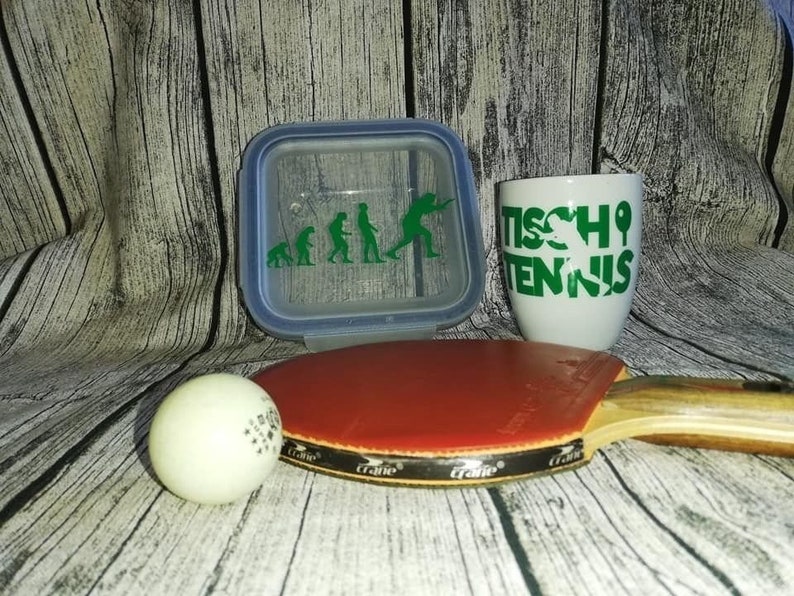 May include: A white ceramic mug with green text that reads "Tischtennis" and a green silhouette of a person playing table tennis. A white table tennis ball, a red and black table tennis paddle, and a clear plastic container with a green silhouette of the evolution of humans playing table tennis.