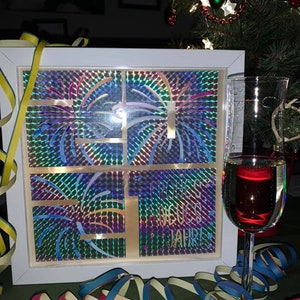 Puede incluir: Una caja enmarcada en blanco con un dise&ntilde;o hologr&aacute;fico de fuegos artificiales. La caja tiene el texto "Frohes Neues Jahr!" en el interior. Un vaso de vino tinto est&aacute; en primer plano con serpentinas de colores.