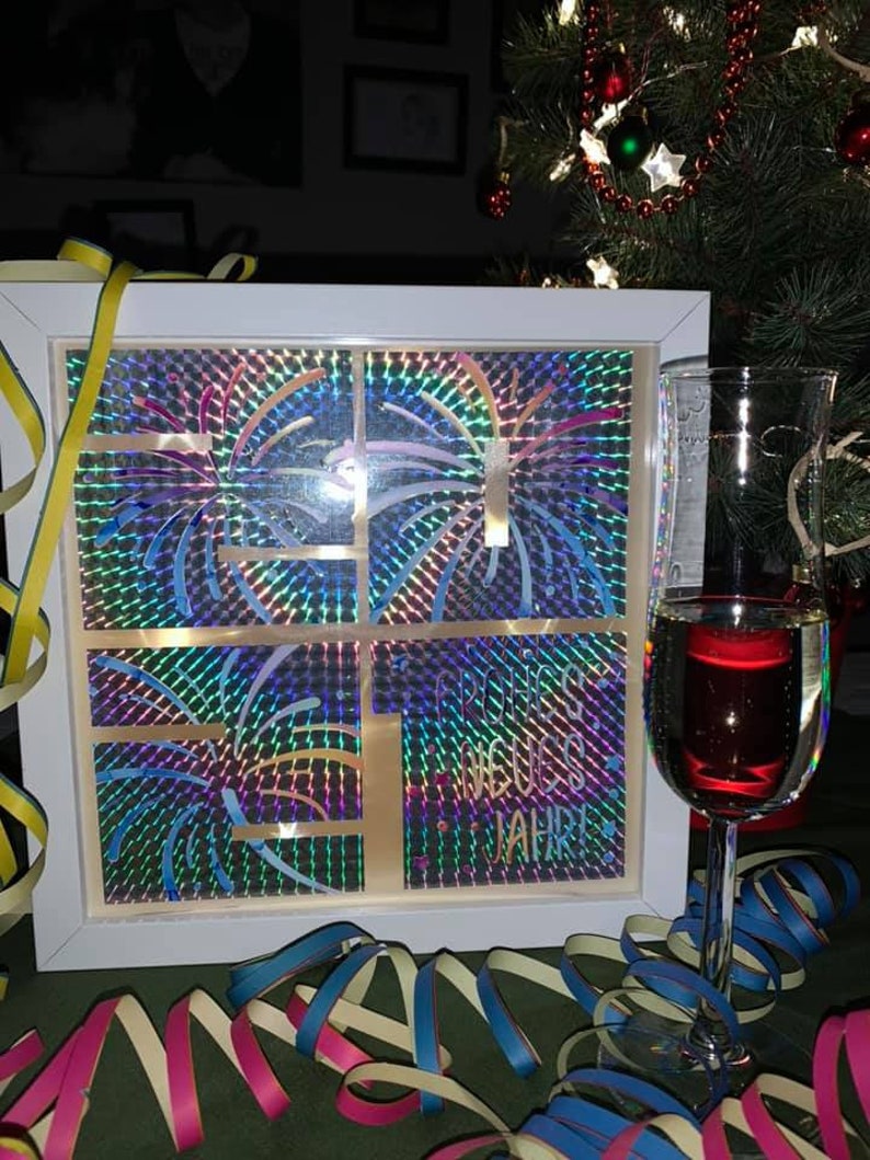 Puede incluir: Una caja enmarcada en blanco con un dise&ntilde;o hologr&aacute;fico de fuegos artificiales. La caja tiene el texto "Frohes Neues Jahr!" en el interior. Un vaso de vino tinto est&aacute; en primer plano con serpentinas de colores.