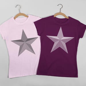 Könnte beinhalten: Zwei T-Shirts, eines in Hellrosa und eines in Dunkelviolett, jeweils mit einem großen, grauen Sterndesign. Die Sterne haben ein genähtes Aussehen, das Textur verleiht. Beide Shirts werden auf Holzbügeln vor einem hellgrauen Hintergrund präsentiert.