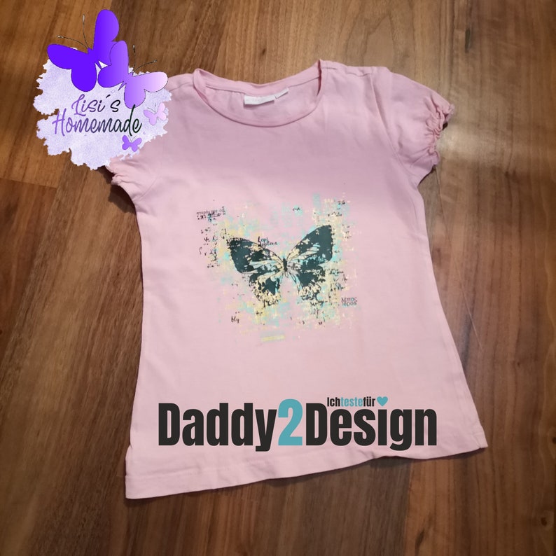 Op de afbeelding: Roze T-shirt met een zwarte vlinderprint en de tekst "Daddy2Design" in blauw en zwart.
