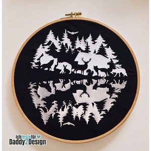 Op de afbeelding: Een zwarte stoffen ring met een wit silhouetontwerp van een bos met dieren en bomen die in een meer worden weerspiegeld. De tekst "WIM" is zichtbaar in de reflectie.