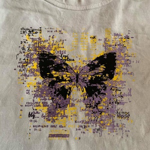Op de afbeelding: Een wit T-shirt met een zwarte en paarse vlinderprint. De vlinder is omringd door gele en paarse tekst, waaronder de woorden "Lepi Hoptera" en "&Alpha;&lambda;&epsilon;&pi;&omicron;&sigmaf; lepos".