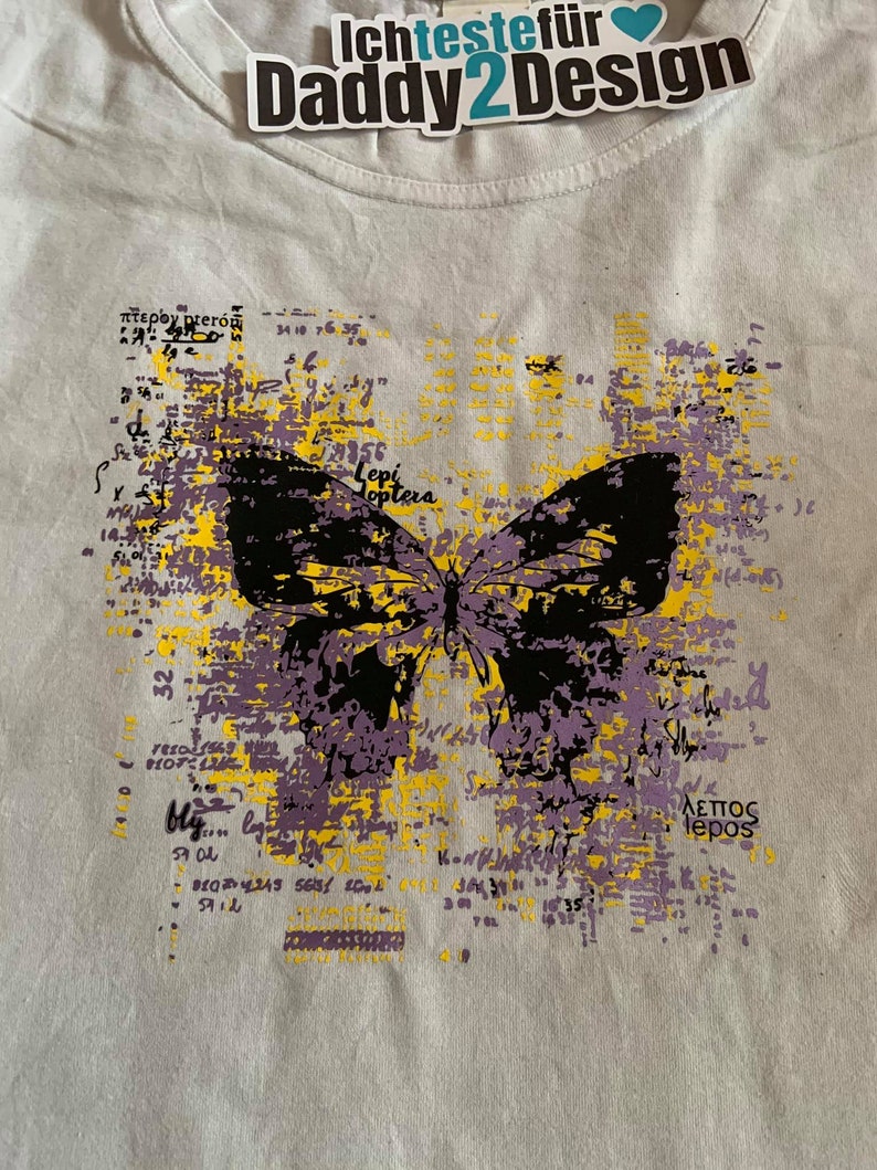 Op de afbeelding: Een wit T-shirt met een zwarte en paarse vlinderprint. De vlinder is omringd door gele en paarse tekst, waaronder de woorden "Lepi Hoptera" en "&Alpha;&lambda;&epsilon;&pi;&omicron;&sigmaf; lepos".