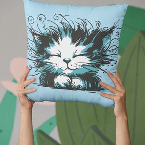 Könnte beinhalten: Hellblaues quadratisches Kissen mit einer Cartoon-Katze mit geschlossenen Augen. Die Katze hat weißes Fell mit schwarzen und türkisfarbenen Akzenten. Das Kissen wird von zwei Händen vor einem grünen und grauen Hintergrund gehalten.