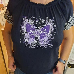 Op de afbeelding: Een donkerblauw shirt met een paarse vlinderprint. Het shirt heeft een gerimpelde halslijn en witte kantversiering op de mouwen.