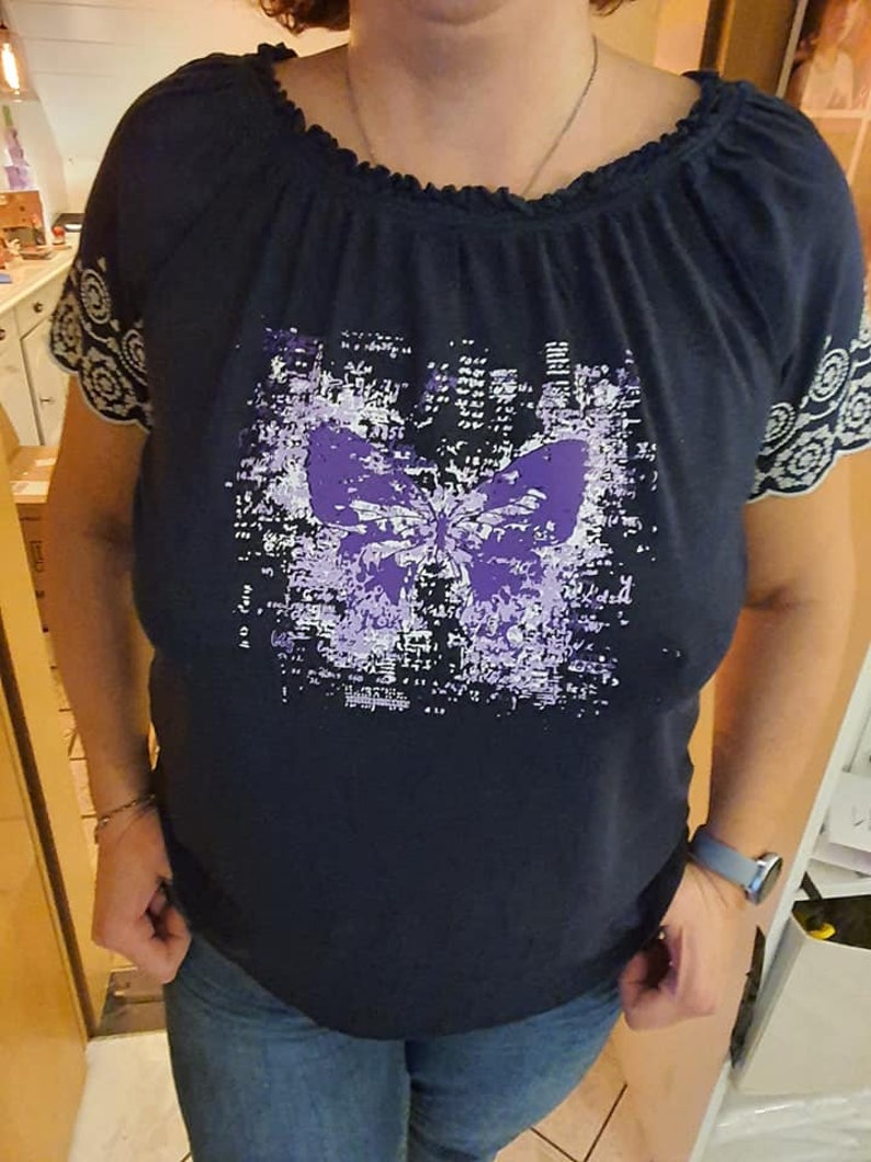 Op de afbeelding: Een donkerblauw shirt met een paarse vlinderprint. Het shirt heeft een gerimpelde halslijn en witte kantversiering op de mouwen.