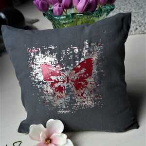 Op de afbeelding: Een grijs kussen met een zilveren en rood vlinderontwerp. Het kussen heeft een verweerde look en is versierd met een witte bloem.
