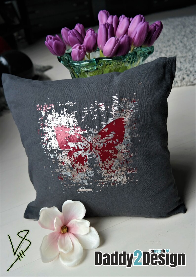 Op de afbeelding: Een grijs kussen met een zilveren en rood vlinderontwerp. Het kussen heeft een verweerde look en is versierd met een witte bloem.