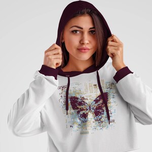 Op de afbeelding: Witte getailleerde hoodie met een bordeauxrode capuchon en manchetten. De hoodie heeft een afbeelding van een vlinder met een blauw en gouden patroon op een witte achtergrond met een textuurontwerp.
