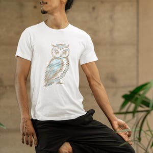 Könnte beinhalten: Weißes T-Shirt mit einem Eulen-Design aus kleinen Punkten in Braun, Blau und Weiß. Das T-Shirt hat einen Rundhalsausschnitt und kurze Ärmel. Die Person trägt eine schwarze Hose. Das Eulen-Design ist der Blickfang.