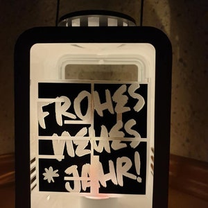 Puede incluir: Una linterna de metal negro con un panel de vidrio esmerilado blanco. El panel de vidrio tiene una pegatina negra con las palabras "Frohes Neues Jahr!" escritas en blanco.