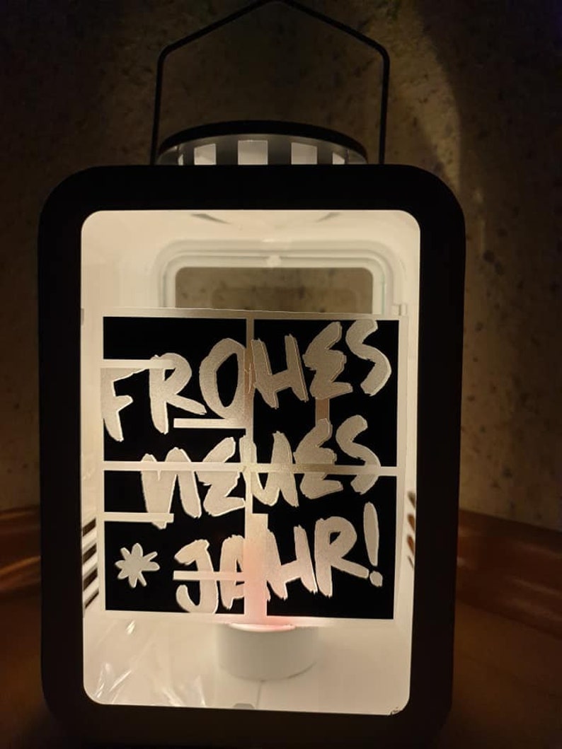 Puede incluir: Una linterna de metal negro con un panel de vidrio esmerilado blanco. El panel de vidrio tiene una pegatina negra con las palabras "Frohes Neues Jahr!" escritas en blanco.